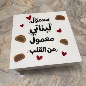 May include: A white box with Arabic text and decorative elements. The text includes the words "maamoul" and "Lebanese." Red heart shapes and small brown cookie illustrations are scattered across the box's surface.