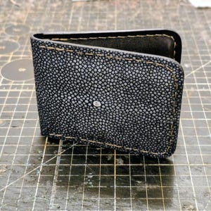 May include: A bifold wallet crafted from dark grey stingray leather featuring a distinctive pebbled texture and a single white circular mark in the centre. The edges are finished with thick tan stitching, and the interior is lined with black leather.