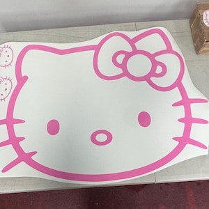 May include: A large white vinyl decal featuring the pink outline of the Hello Kitty character face, including the signature bow, oval eyes, nose, and whiskers. Two smaller versions of the same Hello Kitty face are printed on the left side.