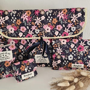 Könnte beinhalten: Ein Set von Accessoires mit floralem Print in Marineblau, Rosa und Pfirsich. Das Set umfasst eine Wickelunterlage, ein kleines Portemonnaie und kleine Beutel. Die Texte "Douceur de vie" und "Bonjour" sind auf den Artikeln zu sehen.