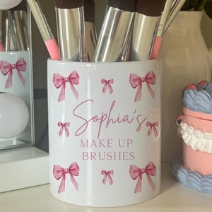 May include: White ceramic makeup brush holder with pink bows and the text "Sophia's Make Up Brushes". The holder is filled with a variety of makeup brushes.