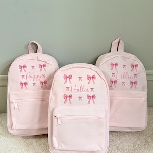 Puede incluir: Tres mochilas de color rosa claro con bolsillos delanteros con cremallera y asas superiores. Cada mochila presenta un nombre en fuente cursiva y gr&aacute;ficos de lazos rosas. Los nombres son "Poppy", "Hallie" y "Lillie".