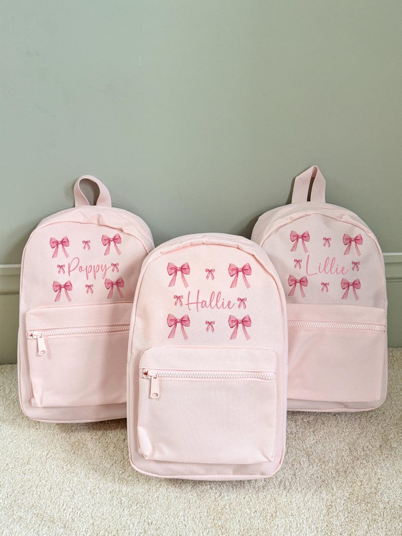 Puede incluir: Tres mochilas de color rosa claro con bolsillos delanteros con cremallera y asas superiores. Cada mochila presenta un nombre en fuente cursiva y gr&aacute;ficos de lazos rosas. Los nombres son "Poppy", "Hallie" y "Lillie".
