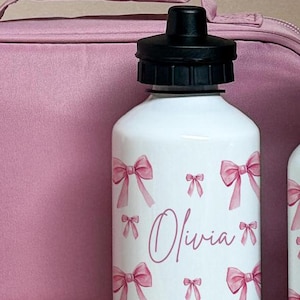 Puede incluir: Botella de agua blanca con tapa negra, decorada con lazos rosas y el nombre "Olivia" en cursiva. Una bolsa de almuerzo rosa se encuentra en el fondo.