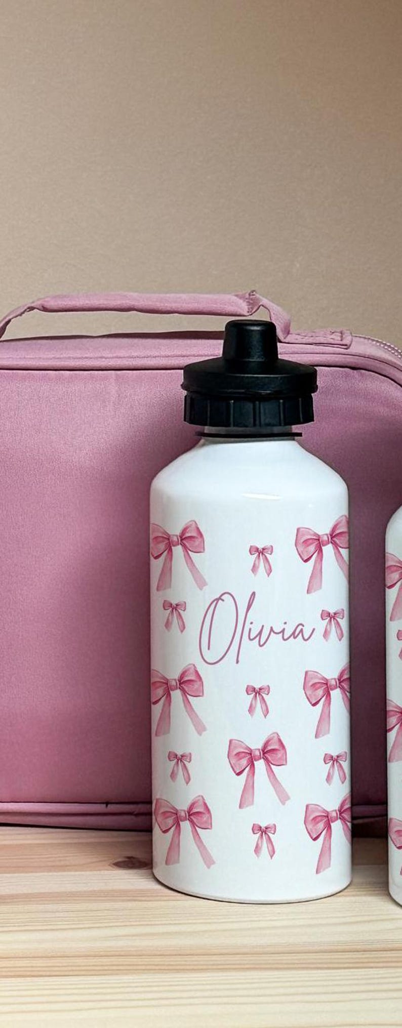 Puede incluir: Botella de agua blanca con tapa negra, decorada con lazos rosas y el nombre "Olivia" en cursiva. Una bolsa de almuerzo rosa se encuentra en el fondo.