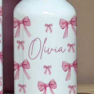 Puede incluir: Botella de agua blanca decorada con lazos rosas y el nombre "Olivia" en cursiva. La botella tiene una tapa negra y est&aacute; sobre una superficie de madera. Una bolsa rosa es parcialmente visible en el fondo.