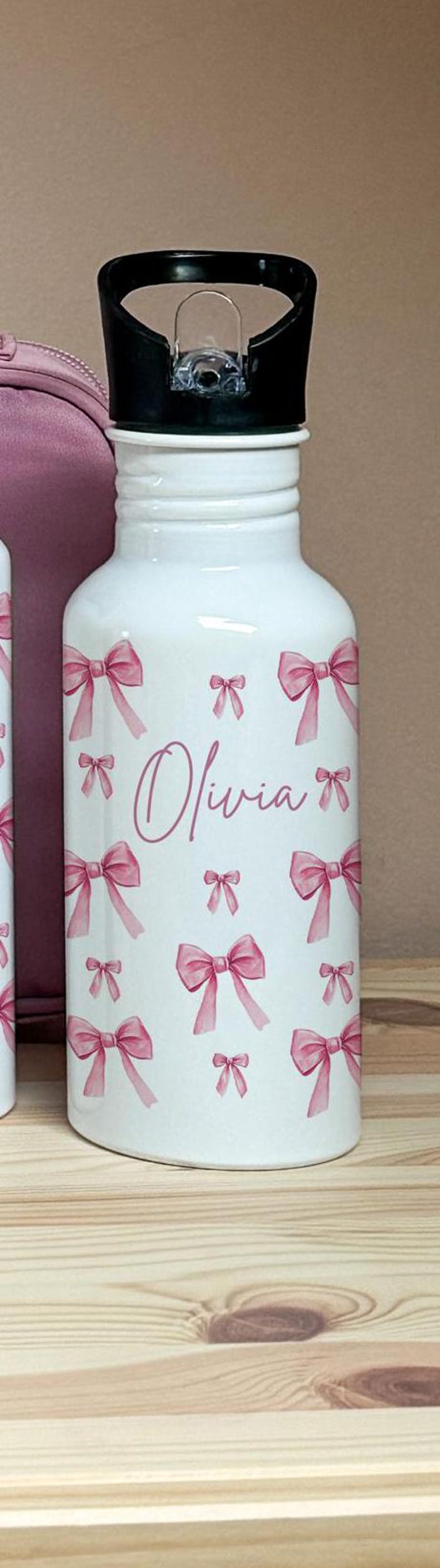 Puede incluir: Botella de agua blanca decorada con lazos rosas y el nombre "Olivia" en cursiva. La botella tiene una tapa negra y est&aacute; sobre una superficie de madera. Una bolsa rosa es parcialmente visible en el fondo.