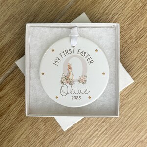Może przedstawiać: Biała ceramiczna ozdoba z złotym kr&oacute;likiem i kwiatowym wzorem. Ozdoba zawiera napis "My First Easter" i jest spersonalizowana imieniem "Olive" i rokiem "2025".