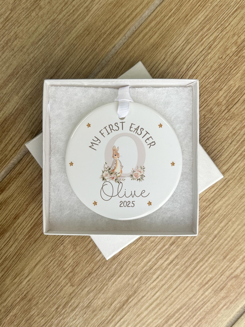 Może przedstawiać: Biała ceramiczna ozdoba z złotym kr&oacute;likiem i kwiatowym wzorem. Ozdoba zawiera napis "My First Easter" i jest spersonalizowana imieniem "Olive" i rokiem "2025".