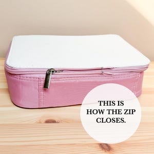 Puede incluir: Un estuche rectangular rosa con una parte superior blanca y un cierre de cremallera plateado. El texto "THIS IS HOW THE ZIP CLOSES" est&aacute; en un c&iacute;rculo blanco en el lado derecho de la imagen.
