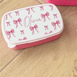Puede incluir: Una fiambrera rectangular rosa con una tapa blanca con un estampado de lazos rosas. El nombre "Olivia" est&aacute; escrito en cursiva en la tapa. La fiambrera est&aacute; sobre una superficie de madera.