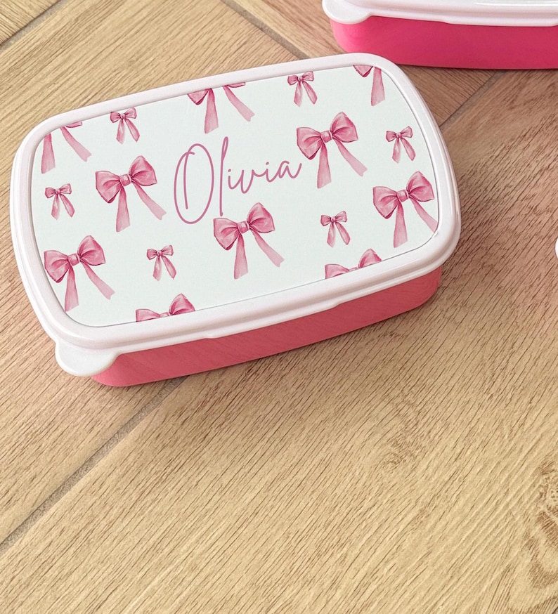 Puede incluir: Una fiambrera rectangular rosa con una tapa blanca con un estampado de lazos rosas. El nombre "Olivia" est&aacute; escrito en cursiva en la tapa. La fiambrera est&aacute; sobre una superficie de madera.