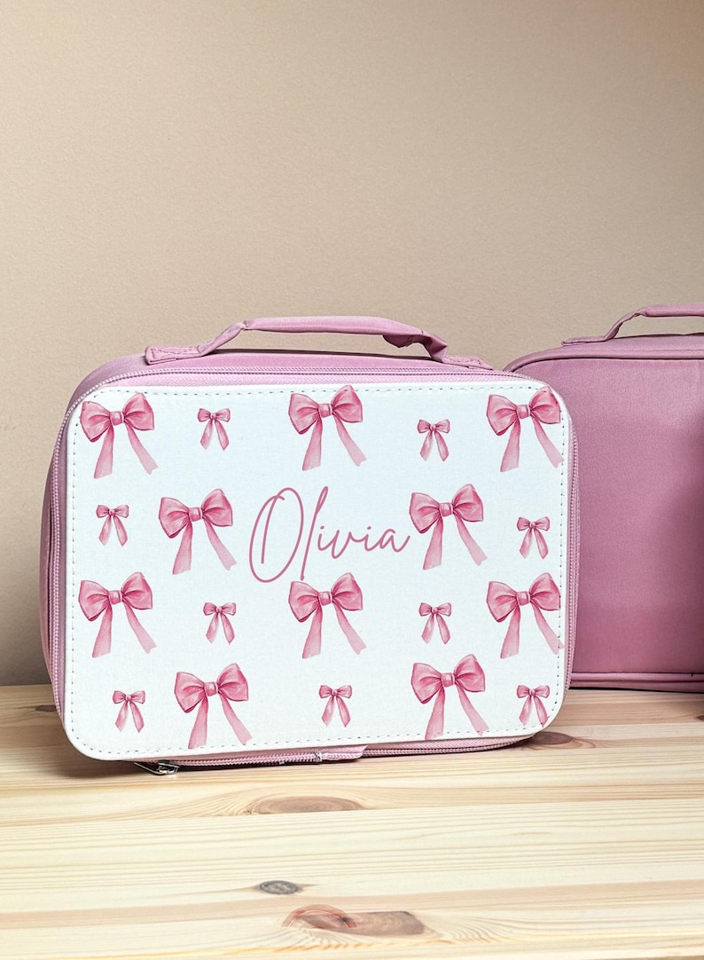 Puede incluir: Bolsa de almuerzo rosa con un panel frontal blanco con un patr&oacute;n de lazos rosas. El nombre "Olivia" est&aacute; escrito en cursiva. Una segunda bolsa rosa s&oacute;lida es visible en el fondo. La bolsa tiene un asa.