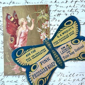May include: A rectangular card featuring a fairy with large butterfly wings and a pink dress, alongside a butterfly-shaped advertisement for G.A. Wrisley's Pink Prussian Soap, which is a laundry and toilet soap combined, both resting on handwritten script.