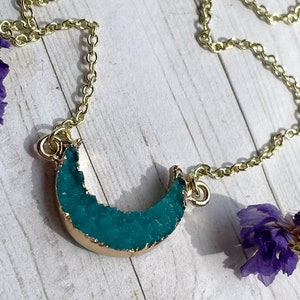 May include: A gold chain necklace with a teal blue crescent moon pendant. The pendant is made of a crystal-like material.