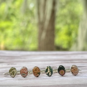 May include: Six gemstone rings with oval-shaped stones in various colors, including green, pink, and brown, are displayed on a wooden surface. Each ring has a silver band and a decorative bezel setting. The background is a blurred view of green foliage.