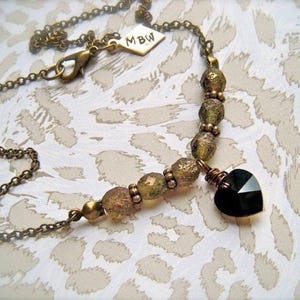 May include: A bronze chain necklace with a black heart pendant and gold and green beads. The necklace has a tag with the initials "MBW" on it.