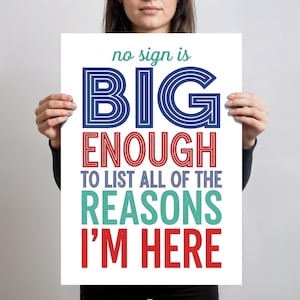 Puede incluir: Un póster blanco con el texto "no sign is BIG ENOUGH TO LIST ALL OF THE REASONS I'M HERE" en letras azules, rojas y turquesas. El póster está sostenido por una persona.