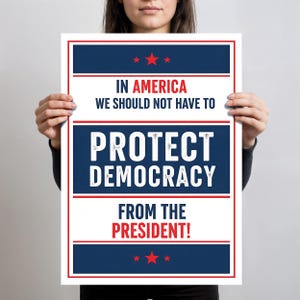 May include: A poster with a white border and a navy blue and red design. The text reads "IN AMERICA WE SHOULD NOT HAVE TO PROTECT DEMOCRACY FROM THE PRESIDENT!" with red stars at the top and bottom.