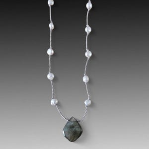 May include: A silver necklace featuring a series of white pearls strung along a delicate chain. The necklace culminates in a faceted, teardrop-shaped, gray-green gemstone pendant. The design is simple and elegant.
