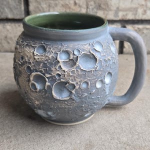 Puede incluir: Un juego de tres tazas de cerámica con acabado mate y moteado. Las tazas son de color crema, gris claro y gris oscuro. Cada taza tiene un diseño simple y minimalista.