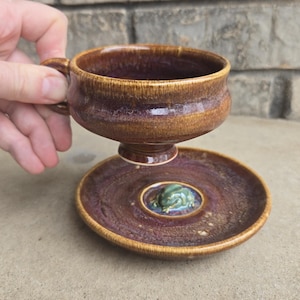 May include: A handmade ceramic teacup and saucer set. The cup and saucer are brown and purple with a glossy finish. The saucer has a raised center with a green and blue design. The cup is held by a hand.