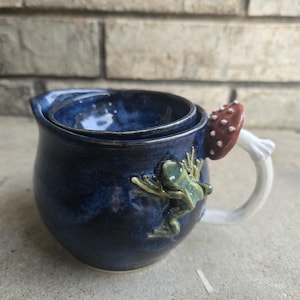 Puede incluir: Taza de cerámica hecha a mano con un esmalte azul oscuro. La taza presenta una rana trepando por el lateral y un asa de seta. La seta es roja con puntos blancos. La taza mide aproximadamente 10 cm de altura.