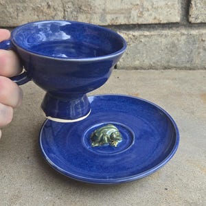 May include: A handmade, cobalt blue ceramic teacup and saucer set. The teacup has a pedestal base and a handle. The saucer features a small, green frog figurine in the centre. The set is displayed on a concrete surface.