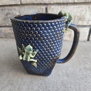 Puede incluir: Taza de cerámica azul oscuro con superficie texturizada y asa marrón. Dos figuras de rana verde claro están adheridas, una en el borde y otra en el lateral. La taza tiene un aspecto artesanal y es adecuada para bebidas calientes.