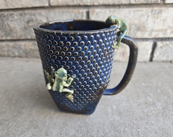 May include: A ceramic mug with a textured, dark blue exterior and a brown handle. Two light green frog figurines are attached to the mug, one climbing the rim and the other on the side. The mug has a unique, handcrafted appearance.