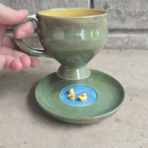 May include: A handmade ceramic teacup and saucer set. The cup and saucer are green with a yellow interior. The saucer features a blue pond design with two yellow duck figurines. The cup has a handle.