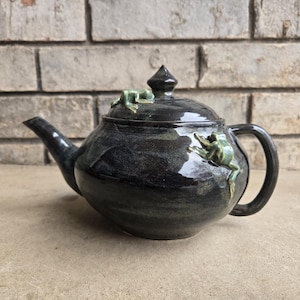 May include: A dark, ceramic teapot with a glossy finish. The teapot features a curved spout, a rounded body, and a handle. Two green frog figurines are attached to the teapot, one on the lid and one on the side. The teapot is sitting on a light-colored surface.