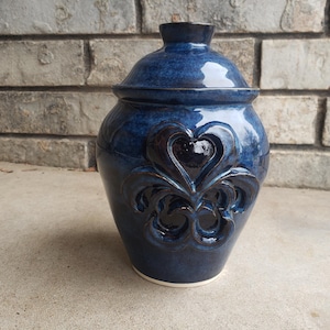 Könnte beinhalten: Eine blaue Keramikdose mit Deckel und einem dekorativen Fleur-de-lis-Design. Die Dose hat eine dunkelblaue Glasur mit einem dezenten, gesprenkelten Effekt.
