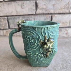 Puede incluir: Una taza de cerámica verde azulado con asa, con diseños florales y de uvas en relieve. Dos pequeñas figuras de rana están adheridas a la taza, una en el asa y otra en el lateral. La taza tiene un diseño artístico único.