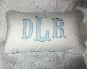 May include: A white pillow with a light blue border and the letters "DLR" embroidered in light blue.