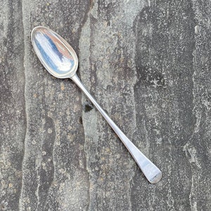 May include: A silver teaspoon with a rounded bowl and a long, slender handle. The spoon is lying on a textured, gray surface.