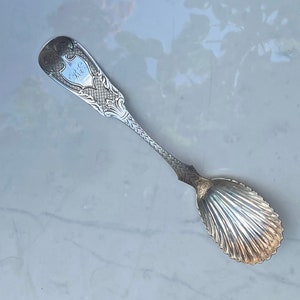 May include: A silver sugar spoon with an ornate handle and a shell-shaped bowl. The handle has an engraved inscription that reads "E.G. Hill".