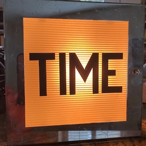 Puede incluir: Un letrero con marco plateado con fondo amarillo y la palabra "TIME" en letras negras. El letrero está iluminado desde atrás, creando un brillo cálido.