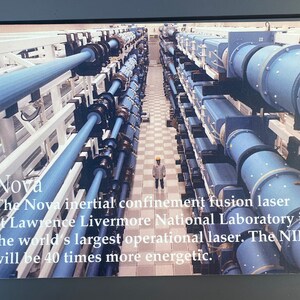 Puede incluir: Imagen del láser de fusión por confinamiento inercial Nova en el Laboratorio Nacional Lawrence Livermore. La imagen muestra un largo pasillo con estructuras cilíndricas azules y una persona con un casco amarillo. El texto indica que es el láser operativo más grande del mundo.