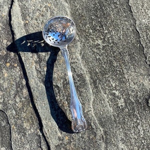 May include: A silver sugar spoon with a decorative, perforated bowl. The spoon is lying on a gray, textured surface.