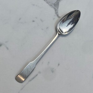 May include: A silver spoon with a rounded bowl and a long, slender handle. The handle has a decorative design.