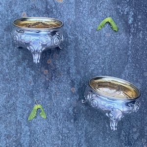 May include: Two silver bowls with gold interiors and ornate designs on a blue textured surface. The bowls have three legs each and are arranged symmetrically.