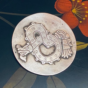 May include: A silver pendant with a detailed design of a serpent with a feathered headdress. The pendant is circular and has a smooth, slightly textured surface.