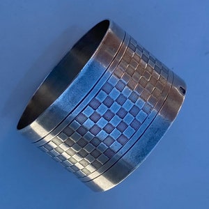 May include: Silver napkin ring with a geometric pattern. The ring is oval shaped and has a smooth, polished finish.