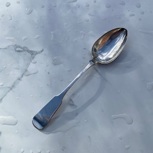 May include: A silver spoon with a rounded bowl and a long, slender handle. The spoon is lying on a white marble surface with water droplets.