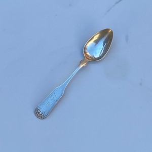 May include: A silver teaspoon with an ornate handle and a rounded bowl. The spoon is on a white marble surface.
