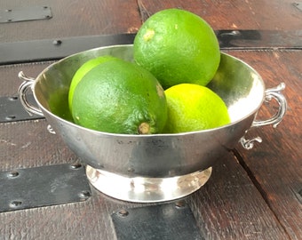 Może przedstawiać: Srebrna miska z czterema zielonymi limonkami w środku. Miska ma dwa uchwyty i stoi na ciemnym drewnianym blacie.