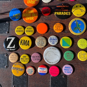 May include: A collection of vintage button pins with various designs and slogans. The pins are mostly round and feature bright colors, including red, yellow, green, blue, and black. Some of the slogans include "I Feel Good All Over", "Love It", "Acquit Socrates", "KMA", "Frodo Lives", "Come to Middle Earth", "When Do We Eat", "Support Your Local Hobbit", and "Marcel Proust is a Yenta".