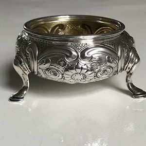 May include: A silver salt cellar with a round, ornate design and three legs. The cellar has a textured surface and a floral pattern.