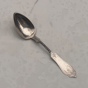 May include: A silver spoon with a twisted handle and an ornate design on the back. The spoon has a pointed oval bowl.
